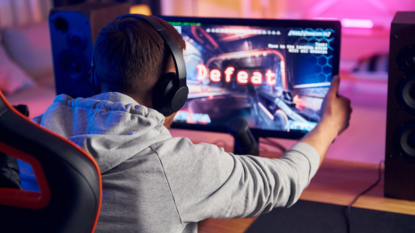 A player in a grey hoodie and headphones, seen from behind, gripping the edge of a monitor that shows the word Defeat over a first-person shooter.