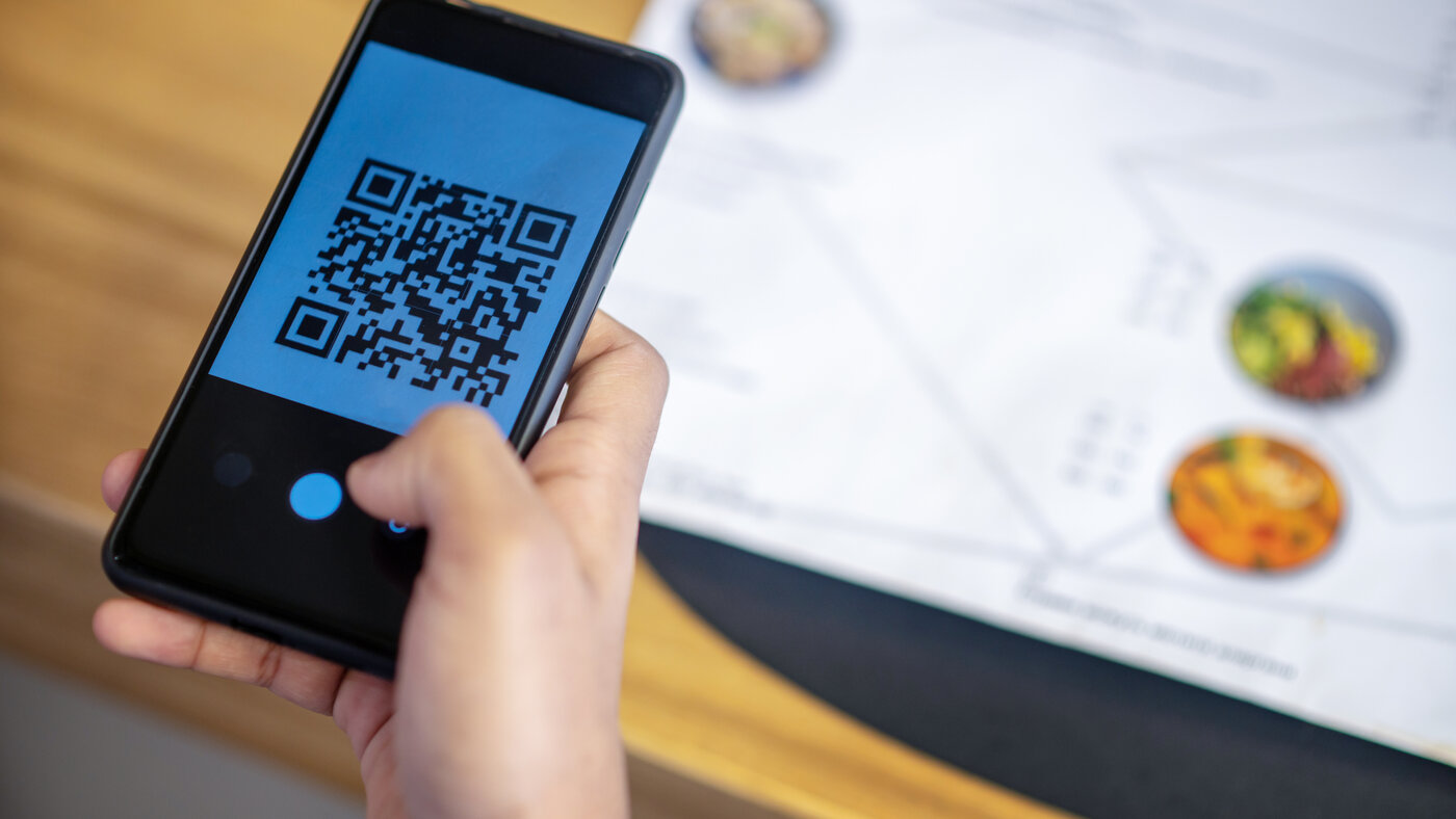 A hand holding a phone whose camera is framing a QR code, with a printed restaurant menu on the table behind it
